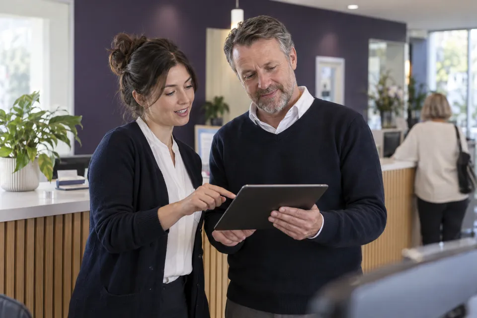 Illustrative scene of two service colleagues coordinating customer arrivals on a tablet behind a reception counter.